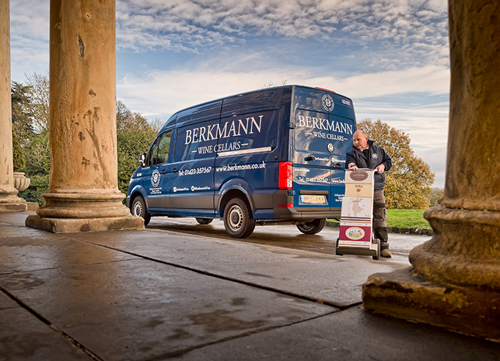 A Berkmann wine delivery van parked outside a large building with a delivery man using a trolley to unload wine.