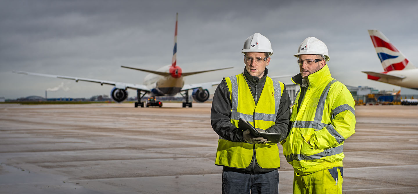 Two workers in yellow Hi-Viz clothing at an airport with two British Airways planes in the background.