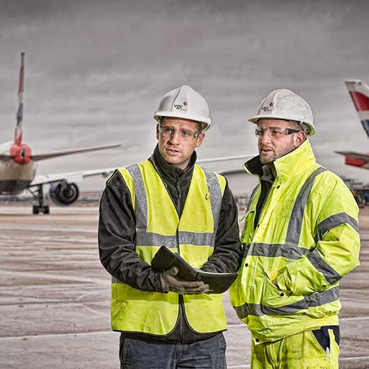 Two workers in hard hats and Hi Viz at Heathrow airport with airoplanes in the background