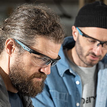 Two carpenters in a workshop discussing a task wearing safety glasses